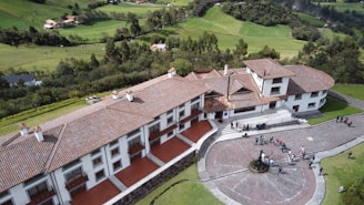 A large, sprawling building with a red tiled roof situated in a lush green landscape. The structure features multiple wings and several chimneys, surrounded by a circular paved area with people gathered around. The grassy hills and trees in the background contribute to a serene and picturesque setting.