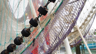 A close-up of a fishing net with various colored ropes and chains interwoven. The net is attached to a series of black, cylindrical floats connected by metal chains. The background includes a partial view of a harbor and a boat, with some structural poles supporting the net.