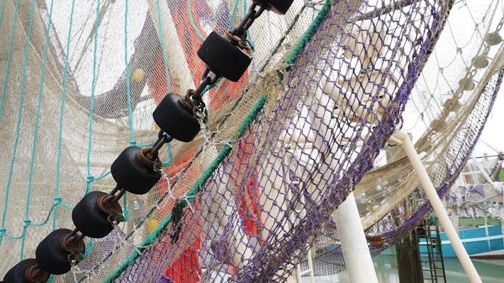 A close-up of a fishing net with various colored ropes and chains interwoven. The net is attached to a series of black, cylindrical floats connected by metal chains. The background includes a partial view of a harbor and a boat, with some structural poles supporting the net.