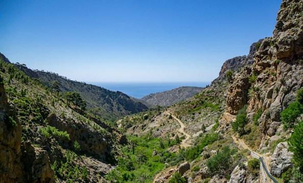 A picturesque landscape featuring a rugged mountainous valley with sparse vegetation. The horizon reveals the blue sea, and the sky is clear with no visible clouds. The terrain is rocky and uneven, with pathways meandering through the greenery.