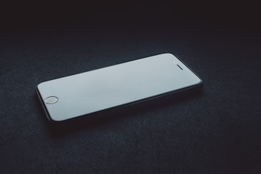 A smartphone with a white screen lies on a dark surface. The lighting casts subtle shadows, giving an emphasis on the phone's sleek design.