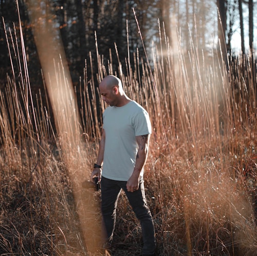 A person stands amidst tall, dry grasses in a sunlit area. The individual is wearing a white shirt and dark pants, holding a camera in one hand. The background is composed of a forest with sunlight filtering through the trees, creating a serene atmosphere.