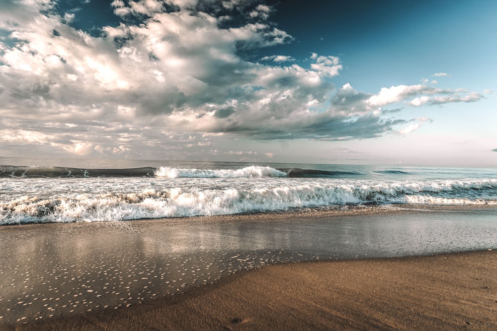ondas do mar batendo na costa sob o céu nublado azul e branco durante o dia