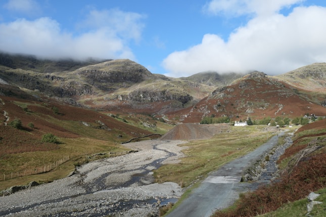 A scenic mountainous landscape with rolling hills and distant peaks partially covered by clouds. A winding road leads through lush greenery with brown patches, likely due to autumn foliage. In the distance, a few small white houses are nestled against the hills.