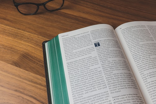 An open book on a wooden table with text visible on its pages. Next to the book, a pair of black eyeglasses is resting on the table surface. The book appears to have green-edged pages.