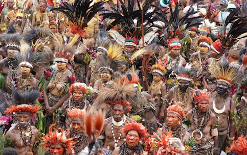 A large group of people adorned in vibrant traditional attire, including elaborate feathered headdresses and face paint. The gathering has a festive and colorful atmosphere, with intricate beadwork and natural materials incorporated into their ceremonial outfits.