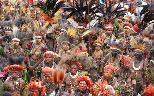 A large group of people adorned in vibrant traditional attire, including elaborate feathered headdresses and face paint. The gathering has a festive and colorful atmosphere, with intricate beadwork and natural materials incorporated into their ceremonial outfits.