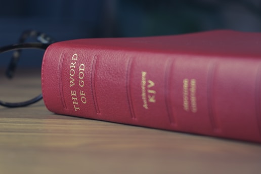 A softly lit open Bible resting on a wooden table, with a pair of reading glasses beside it.
