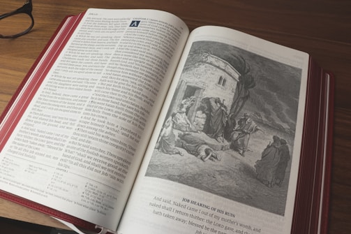 An open book with a color illustration on the right page depicting a historical or biblical scene. The left page contains text in a structured format with columns. The book is lying on a wooden surface, and a pair of glasses is placed to the side.