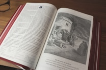 An open book with a color illustration on the right page depicting a historical or biblical scene. The left page contains text in a structured format with columns. The book is lying on a wooden surface, and a pair of glasses is placed to the side.