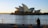 A photographer stands with a tripod on a waterfront capturing the Sydney Opera House at sunrise. The rising sun creates a glare on the iconic sails of the building, casting a warm glow over the scene.