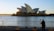 A photographer stands with a tripod on a waterfront capturing the Sydney Opera House at sunrise. The rising sun creates a glare on the iconic sails of the building, casting a warm glow over the scene.
