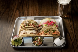 A metal tray holds three tacos, each with different fillings including pickled vegetables, fresh herbs, shredded meat, sliced fruit, and cheese. Accompanying the tacos are small metal cups containing guacamole, salsa, and sour cream. A glass of light beer is placed on the wooden table beside the tray.