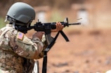 A person wearing a military uniform and helmet aims and fires an assault rifle. An empty shell casing is ejected from the weapon. The uniform features a U.S. flag patch, and the setting appears outdoors with a blurred background.