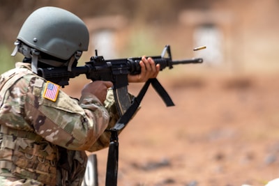 A person wearing a military uniform and helmet aims and fires an assault rifle. An empty shell casing is ejected from the weapon. The uniform features a U.S. flag patch, and the setting appears outdoors with a blurred background.