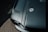 A close-up view of a glossy, dark green car hood featuring a chrome hood pin and cable. The image captures the texture and shine of the surface with light reflecting off the paint. Part of a headlight and the surrounding frame are visible at the bottom left corner.