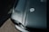 A close-up view of a glossy, dark green car hood featuring a chrome hood pin and cable. The image captures the texture and shine of the surface with light reflecting off the paint. Part of a headlight and the surrounding frame are visible at the bottom left corner.