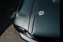 A close-up view of a glossy, dark green car hood featuring a chrome hood pin and cable. The image captures the texture and shine of the surface with light reflecting off the paint. Part of a headlight and the surrounding frame are visible at the bottom left corner.