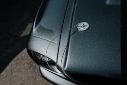 A close-up view of a glossy, dark green car hood featuring a chrome hood pin and cable. The image captures the texture and shine of the surface with light reflecting off the paint. Part of a headlight and the surrounding frame are visible at the bottom left corner.