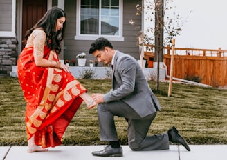 A man dressed in a gray suit is kneeling on a grassy lawn in front of a house, helping a woman adjust her sandal. The woman is wearing a red sari with gold patterns and a cream blouse. The setting includes a wooden fence and some small trees, suggesting a suburban neighborhood.