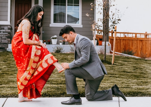 A man dressed in a gray suit is kneeling on a grassy lawn in front of a house, helping a woman adjust her sandal. The woman is wearing a red sari with gold patterns and a cream blouse. The setting includes a wooden fence and some small trees, suggesting a suburban neighborhood.