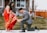 A man dressed in a gray suit is kneeling on a grassy lawn in front of a house, helping a woman adjust her sandal. The woman is wearing a red sari with gold patterns and a cream blouse. The setting includes a wooden fence and some small trees, suggesting a suburban neighborhood.