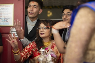 A group of people standing together, appearing engaged in celebration with joyful expressions. The central woman is wearing a traditional red outfit and gold jewelry, holding a ceremonial item. Others around her are dressed in formal attire.