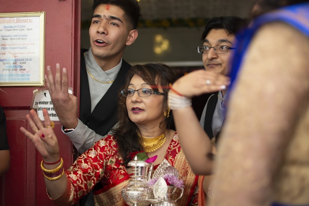 A group of people standing together, appearing engaged in celebration with joyful expressions. The central woman is wearing a traditional red outfit and gold jewelry, holding a ceremonial item. Others around her are dressed in formal attire.