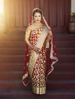 A person is wearing an intricately designed red and gold saree, adorned with gold jewelry. They appear to be standing on steps with wooden doors in the background, and soft sunlight is illuminating the scene.