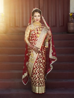 A person is wearing an intricately designed red and gold saree, adorned with gold jewelry. They appear to be standing on steps with wooden doors in the background, and soft sunlight is illuminating the scene.