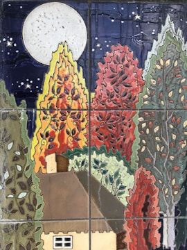 A ceramic tile artwork depicting a whimsical scene of a small house surrounded by colorful and stylized trees. A large full moon shines in the night sky, which is adorned with stars. The trees are vibrantly colored in shades of orange, red, green, and blue, creating an enchanting and lively atmosphere.
