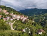 A picturesque hillside town with clustered buildings featuring red and white roofs is nestled amidst lush, green vegetation. The landscape is dominated by rolling hills and a backdrop of forested mountains. A distinct mosque with a tall minaret stands out among the buildings, and a winding road cuts through the greenery leading to the town.