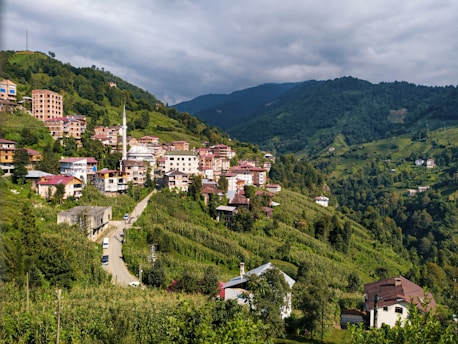A picturesque hillside town with clustered buildings featuring red and white roofs is nestled amidst lush, green vegetation. The landscape is dominated by rolling hills and a backdrop of forested mountains. A distinct mosque with a tall minaret stands out among the buildings, and a winding road cuts through the greenery leading to the town.