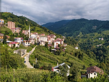 A picturesque hillside town with clustered buildings featuring red and white roofs is nestled amidst lush, green vegetation. The landscape is dominated by rolling hills and a backdrop of forested mountains. A distinct mosque with a tall minaret stands out among the buildings, and a winding road cuts through the greenery leading to the town.