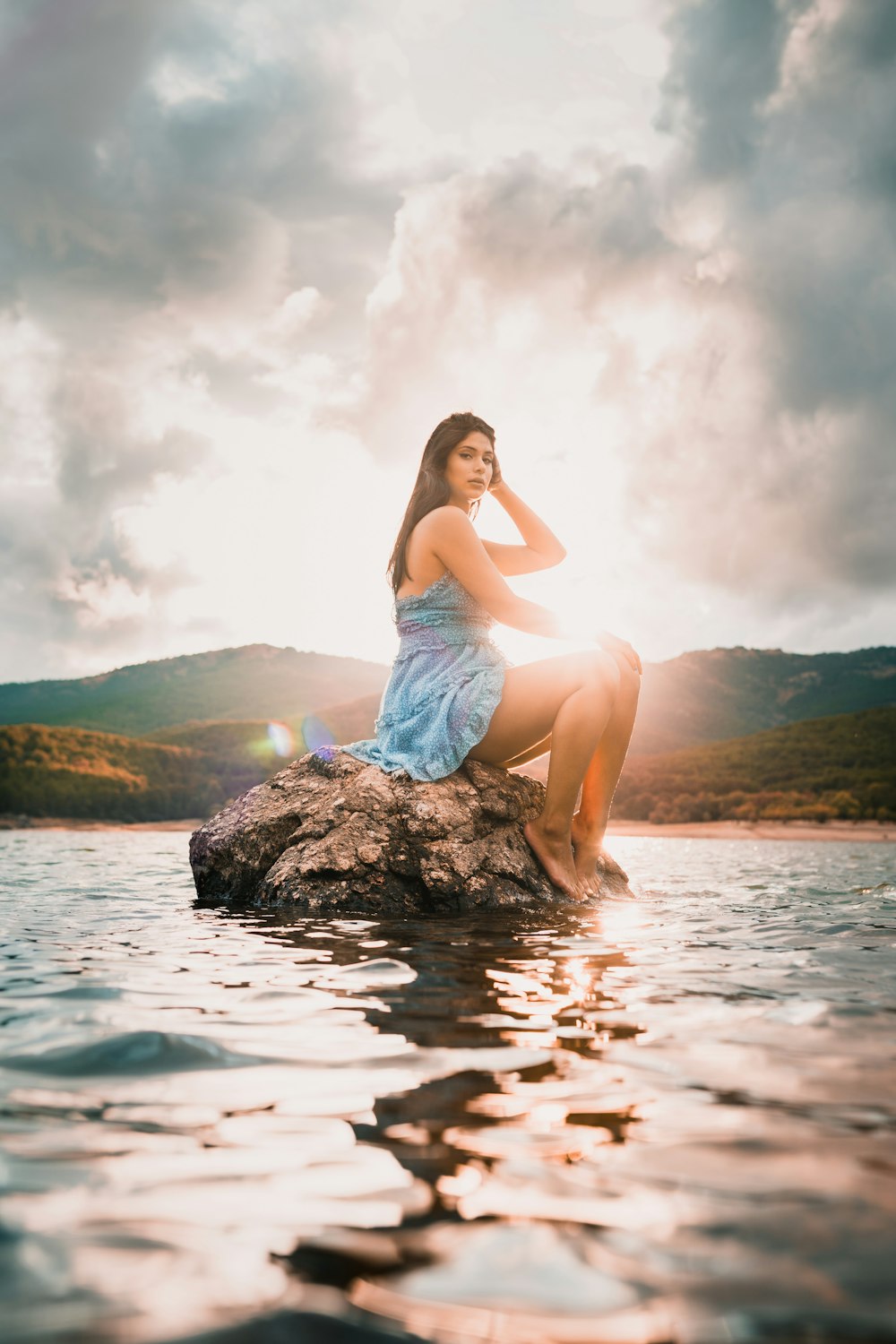woman in white tank top and blue denim shorts sitting on rock in water