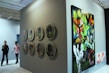 A gallery setting featuring contemporary artwork on display. The left side of the image showcases a series of circular artworks arranged in two vertical rows on a dark wall. To the right, there is a large, colorful painting depicting vibrant and abstract figures and scenes. Two people are walking through the gallery, observing the art pieces.