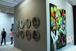 A gallery setting featuring contemporary artwork on display. The left side of the image showcases a series of circular artworks arranged in two vertical rows on a dark wall. To the right, there is a large, colorful painting depicting vibrant and abstract figures and scenes. Two people are walking through the gallery, observing the art pieces.