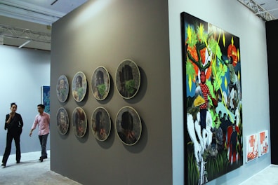 A gallery setting featuring contemporary artwork on display. The left side of the image showcases a series of circular artworks arranged in two vertical rows on a dark wall. To the right, there is a large, colorful painting depicting vibrant and abstract figures and scenes. Two people are walking through the gallery, observing the art pieces.