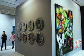 A gallery setting featuring contemporary artwork on display. The left side of the image showcases a series of circular artworks arranged in two vertical rows on a dark wall. To the right, there is a large, colorful painting depicting vibrant and abstract figures and scenes. Two people are walking through the gallery, observing the art pieces.