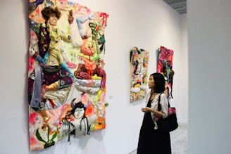 A person observes vibrant textile art pieces displayed on a gallery wall. The artwork consists of multicolored fabrics creatively arranged to form abstract, three-dimensional designs that are both colorful and textured. The gallery space is modern with simple white walls.