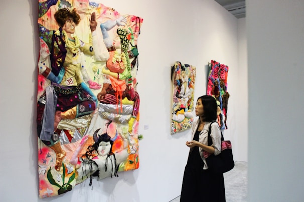 A person observes vibrant textile art pieces displayed on a gallery wall. The artwork consists of multicolored fabrics creatively arranged to form abstract, three-dimensional designs that are both colorful and textured. The gallery space is modern with simple white walls.