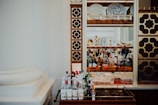 A bar setup featuring a decorative cabinet with intricate brown and gold geometric patterns, housing an assortment of liquor bottles, glassware, and decorative plates. A trolley positioned in the foreground holds various bottles.