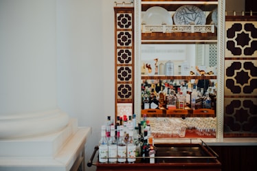 A bar setup featuring a decorative cabinet with intricate brown and gold geometric patterns, housing an assortment of liquor bottles, glassware, and decorative plates. A trolley positioned in the foreground holds various bottles.