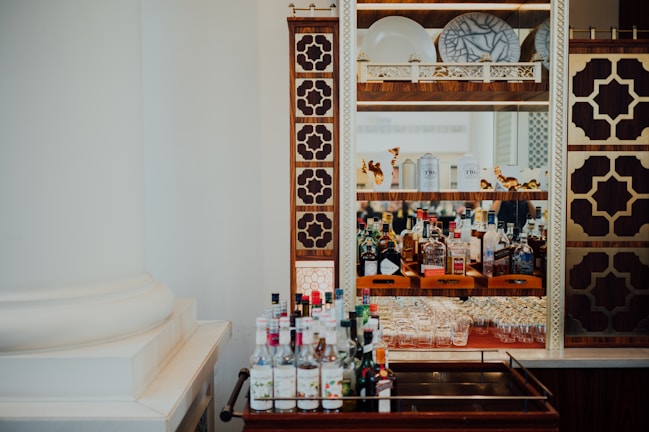 A bar setup featuring a decorative cabinet with intricate brown and gold geometric patterns, housing an assortment of liquor bottles, glassware, and decorative plates. A trolley positioned in the foreground holds various bottles.