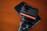 A compact action camera with a lens and screen, mounted on a tripod adapter. The equipment rests on a flat, dark surface, potentially a smartphone or tablet. The background has a warm, wooden texture.