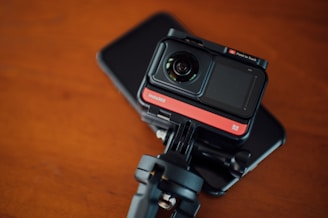 A compact action camera with a lens and screen, mounted on a tripod adapter. The equipment rests on a flat, dark surface, potentially a smartphone or tablet. The background has a warm, wooden texture.