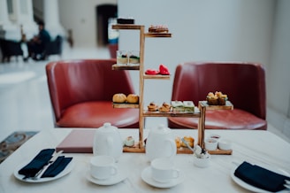 A sophisticated afternoon tea setting with a tiered tray filled with various pastries and desserts is arranged on a marble table. Two white teapots, cups, and saucers accompany the setup, alongside small dishes of condiments. Two red leather chairs are positioned in the background, adding a touch of elegance to the scene. The lighting is soft and natural, giving the space a welcoming ambiance.