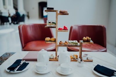 A sophisticated afternoon tea setting with a tiered tray filled with various pastries and desserts is arranged on a marble table. Two white teapots, cups, and saucers accompany the setup, alongside small dishes of condiments. Two red leather chairs are positioned in the background, adding a touch of elegance to the scene. The lighting is soft and natural, giving the space a welcoming ambiance.