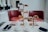 A sophisticated afternoon tea setting with a tiered tray filled with various pastries and desserts is arranged on a marble table. Two white teapots, cups, and saucers accompany the setup, alongside small dishes of condiments. Two red leather chairs are positioned in the background, adding a touch of elegance to the scene. The lighting is soft and natural, giving the space a welcoming ambiance.