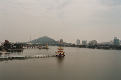 A serene lake with a traditional Chinese pavilion extending into the water. The backdrop features a cityscape with modern buildings and lush greenery. In the distance, a mountain looms under a cloudy sky, creating a peaceful and balanced scene.